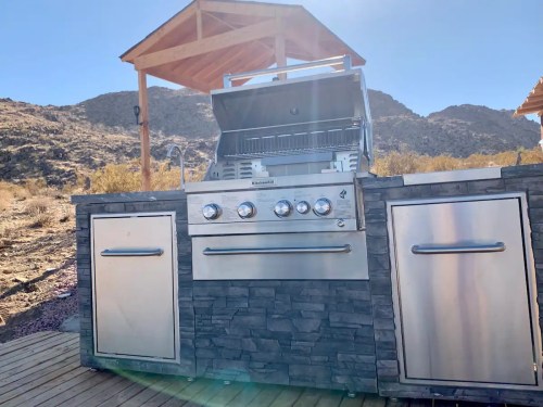 A modern outdoor grill with a stone base, set against a desert landscape and clear blue sky.