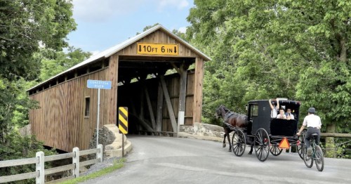 A horse-drawn carriage approaches a wooden covered bridge surrounded by lush greenery.