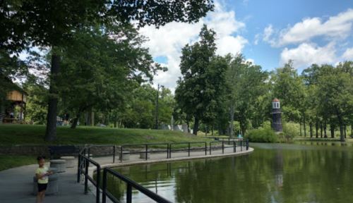 A serene park scene with a pond, trees, and a child standing near the water's edge under a blue sky.