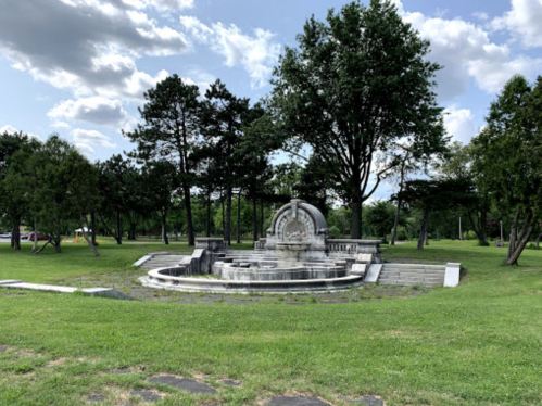 A stone fountain surrounded by grass and trees under a partly cloudy sky.
