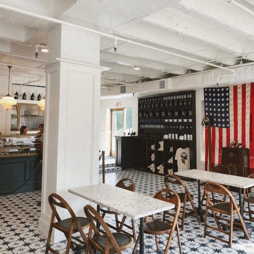 Bright café interior with a large American flag, marble tables, wooden chairs, and patterned tile flooring.