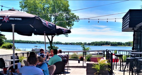 Outdoor dining area by a lake, featuring umbrellas, tables, and patrons enjoying the sunny weather.