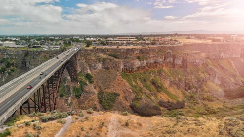 Aerial view of a bridge spanning a canyon, with a town and fields in the background under a cloudy sky.