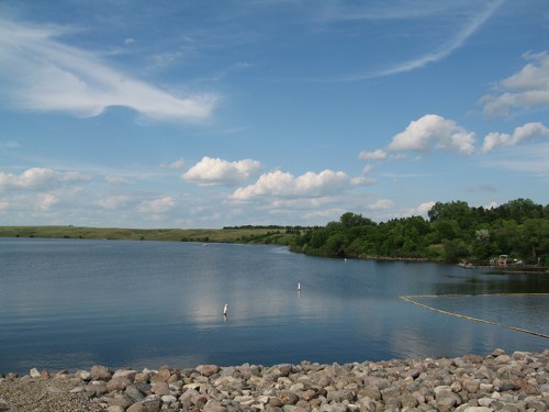 A serene lake surrounded by green hills and a blue sky with fluffy clouds, featuring a rocky shoreline.