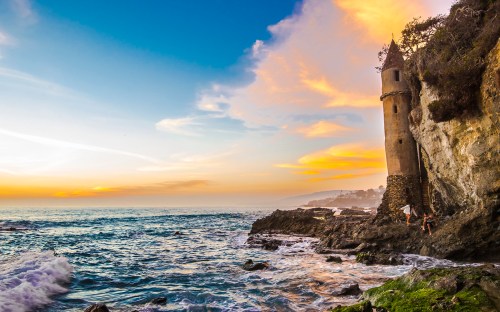 A coastal scene featuring a tower on a rocky shore, with waves crashing and a colorful sunset in the background.
