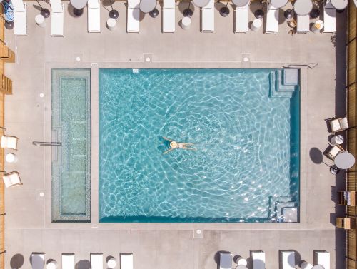 Aerial view of a swimming pool with a person floating, surrounded by lounge chairs and a hot tub.