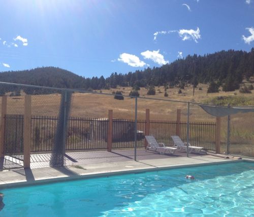 A clear blue pool with lounge chairs, surrounded by a fence and mountains under a bright blue sky with fluffy clouds.
