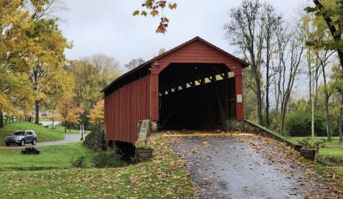 A red covered bridge surrounded by autumn foliage and a gravel path, with a car parked nearby.