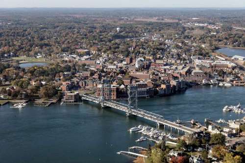 Aerial view of a riverside town with bridges, boats, and autumn foliage in the surrounding landscape.