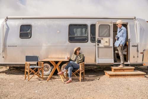 A woman sits at a table outside a silver trailer, while a man stands on the steps, both enjoying a sunny day.