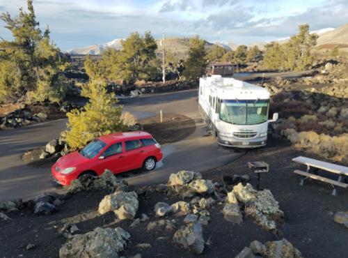 A red car and an RV navigate a winding road through a rocky, volcanic landscape with sparse trees.