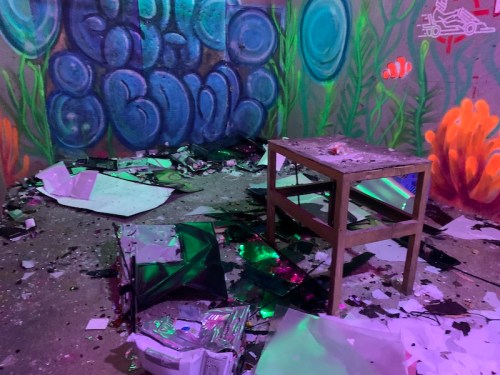 A cluttered room with scattered debris, a wooden table, and colorful graffiti on the walls.