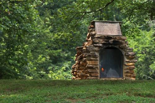 A stone structure with a plaque, surrounded by greenery, set in a peaceful outdoor environment.