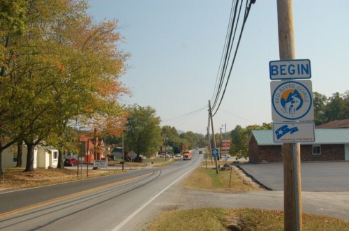 A rural road with a "Begin" sign for the West River Corridor, surrounded by trees and small buildings.