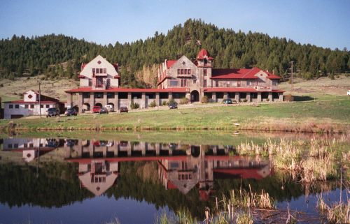 A large, rustic building with red roofs reflects in a calm lake, surrounded by green hills and trees.
