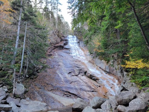 A cascading waterfall flows down rocky terrain, surrounded by lush green trees and autumn foliage.