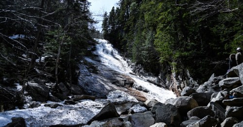 A cascading waterfall flows down rocky terrain, surrounded by lush green trees and patches of snow.
