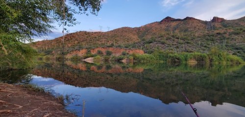 Scenic view of a calm river reflecting mountains and trees under a clear sky, with fishing gear in the foreground.