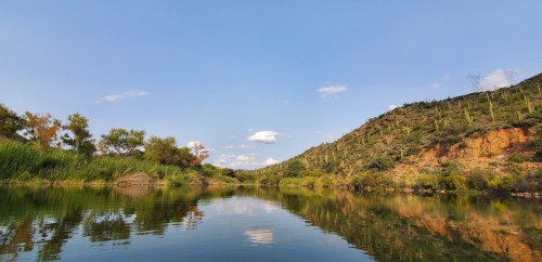 A serene river scene with lush greenery and hills under a clear blue sky, reflecting in the calm water.