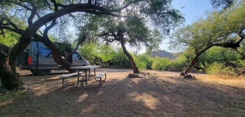 A serene campsite with a picnic table, surrounded by trees and a van parked nearby, under a clear blue sky.