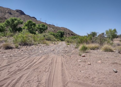 A sandy path leads through sparse vegetation and rocky terrain, with mountains in the background under a clear blue sky.