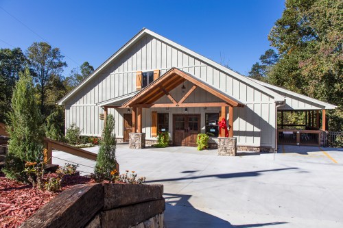 A modern, rustic building with a peaked roof, wooden accents, and a landscaped entrance on a sunny day.
