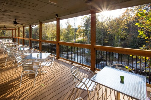 A sunlit outdoor patio with metal tables and chairs overlooking a scenic view of trees and a stream.
