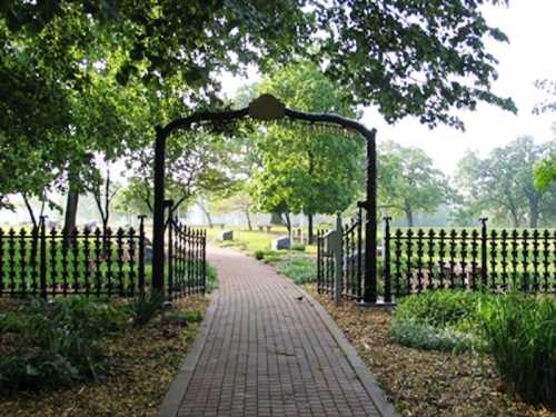 A serene pathway leads through an ornate gate into a lush, green park with trees and gravestones in the background.