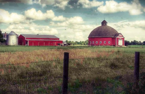 A red barn and silo stand in a grassy field under a cloudy sky, surrounded by a wooden fence.