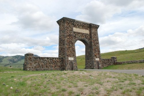 Stone archway with a grassy foreground and mountains in the background under a cloudy sky.