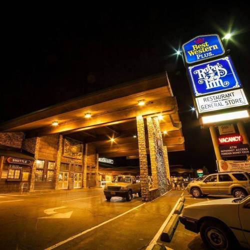 A well-lit hotel entrance at night, featuring a restaurant and general store sign, with parked cars in the foreground.