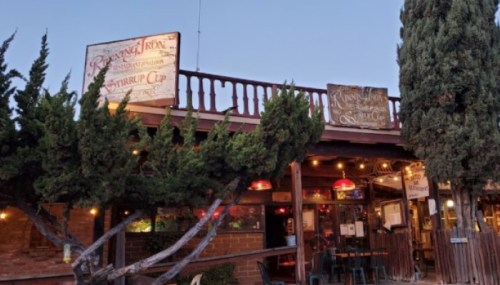 A rustic restaurant exterior with wooden signs, string lights, and trees, set against a twilight sky.