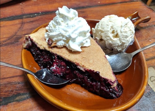 A slice of berry pie topped with whipped cream, served with a scoop of vanilla ice cream on a brown plate.