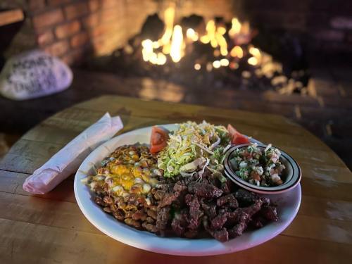 A plate of grilled meat, beans with cheese, salad, and salsa, with a cozy fireplace in the background.