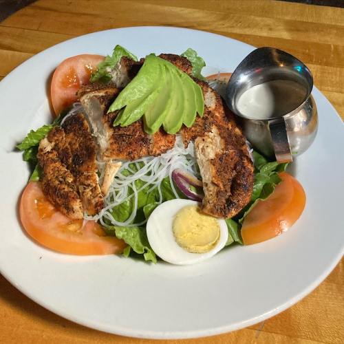 A plate of salad with grilled chicken, avocado, tomatoes, hard-boiled egg, and a small container of dressing.