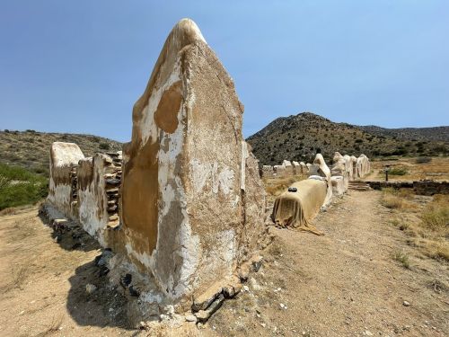 Ruins of weathered stone structures in a dry landscape, with mountains in the background under a clear blue sky.