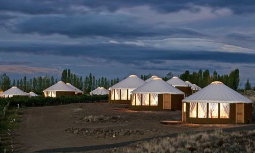 A serene landscape featuring illuminated yurts under a twilight sky, surrounded by trees and open terrain.