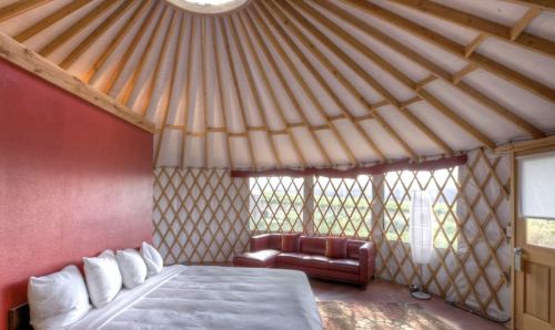 Cozy yurt interior with a circular skylight, wooden beams, a bed, and a red couch against a backdrop of large windows.