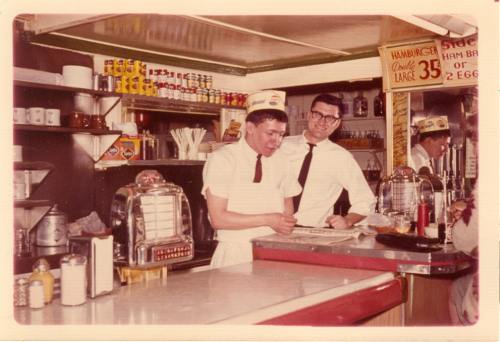 Two men in vintage diner uniforms stand behind a counter, one taking notes, with retro decor and a jukebox in the background.
