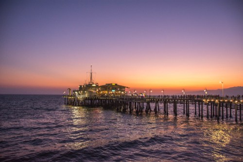 A pier extends into the ocean at sunset, with colorful skies and lights illuminating the structure.