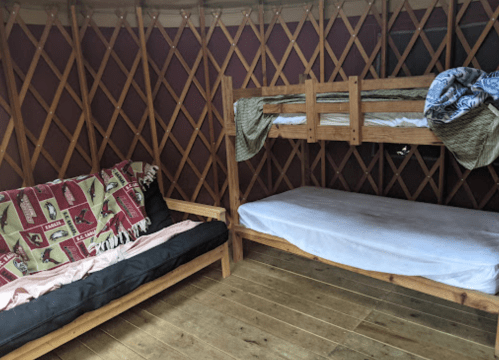 Interior of a yurt featuring a bunk bed and a couch with a patterned blanket. Wooden floor and lattice walls are visible.