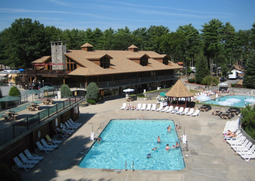 A sunny pool area with lounge chairs, surrounded by trees and a large building in the background. People are swimming and relaxing.