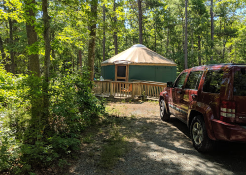 A red SUV parked on a gravel path near a green yurt surrounded by lush trees in a wooded area.
