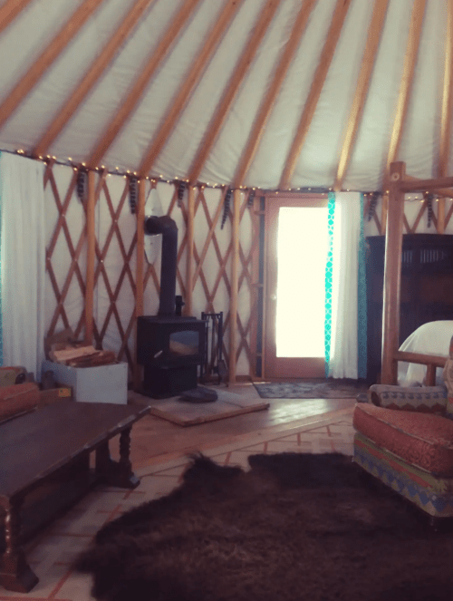 Cozy interior of a yurt with wooden beams, a wood stove, and a door leading outside, featuring a rug and seating area.