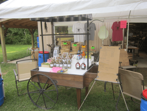 A market cart displaying jars and products, with a tent overhead and a list on a board nearby.