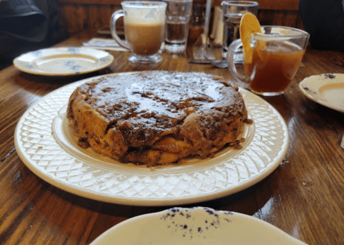 A large, round cinnamon roll pancake on a decorative plate, surrounded by drinks on a wooden table.