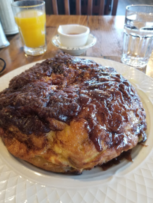 A large, caramelized cinnamon roll on a plate, with glasses of orange juice and water in the background.