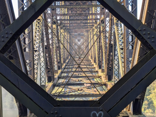 View looking up through a metal bridge structure, showcasing intricate beams and geometric patterns.