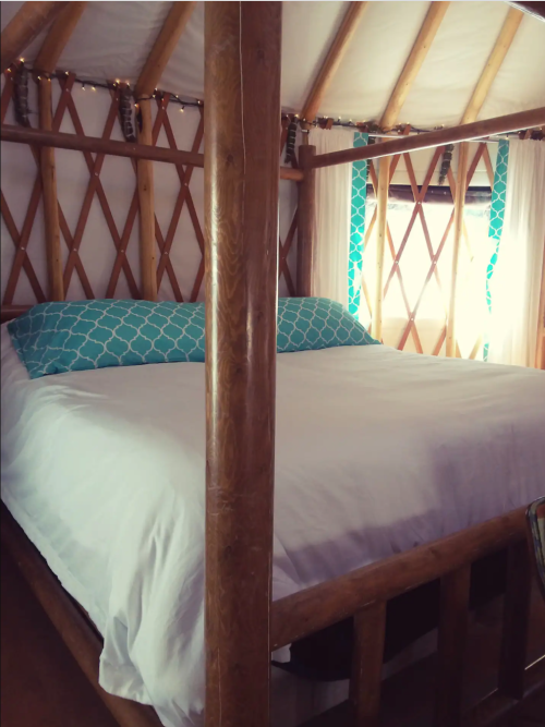 Cozy interior of a yurt featuring a large bed with turquoise pillows and wooden beams. Soft lighting adds warmth.