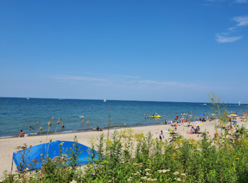 A sunny beach scene with people relaxing, swimming, and a blue tent near the water under a clear blue sky.